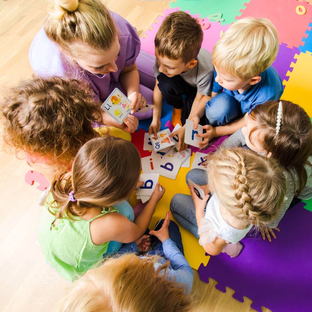 Erzieherin sitzt mit Kindergartenkindern auf dem bunten Spielteppich und liest aus einem Bilderbuch vor. Die Kinder schauen begeistert zu und zeigen auf die Bilder - ein herzlicher Moment pädagogischer Betreuung.