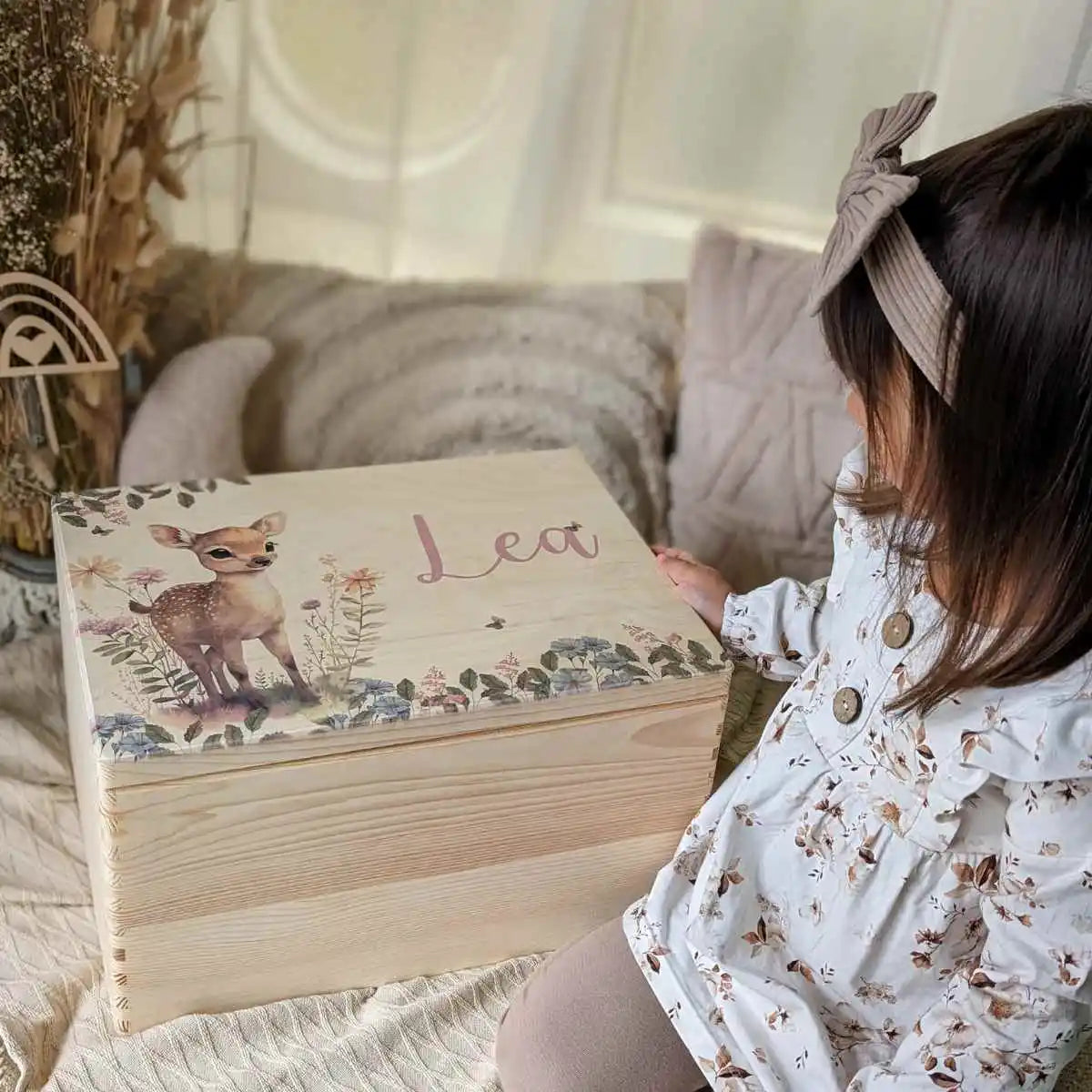 Mädchen mit Haarschleife betrachtet personalisierte Holzbox mit Namen 'Lea' und Rehkitz-Motiv auf weicher Decke im gemütlichen Kinderzimmer mit Naturdeko.