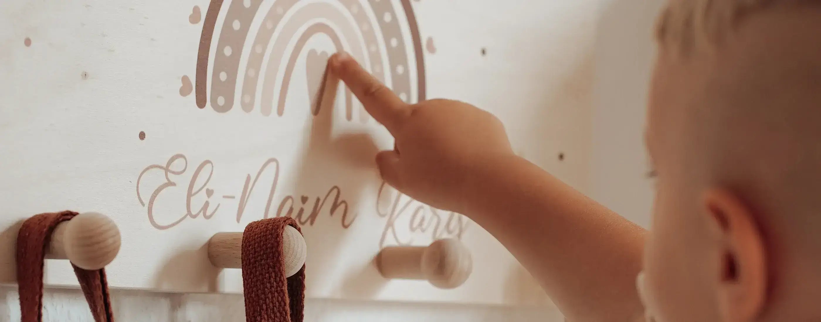 Kinderhand berührt personalisierte Kindergarderobe mit Namen 'Eli-Naim Karo' und Regenbogen-Motiv, Holzhaken mit braunen Bändern für Jacken und Taschen