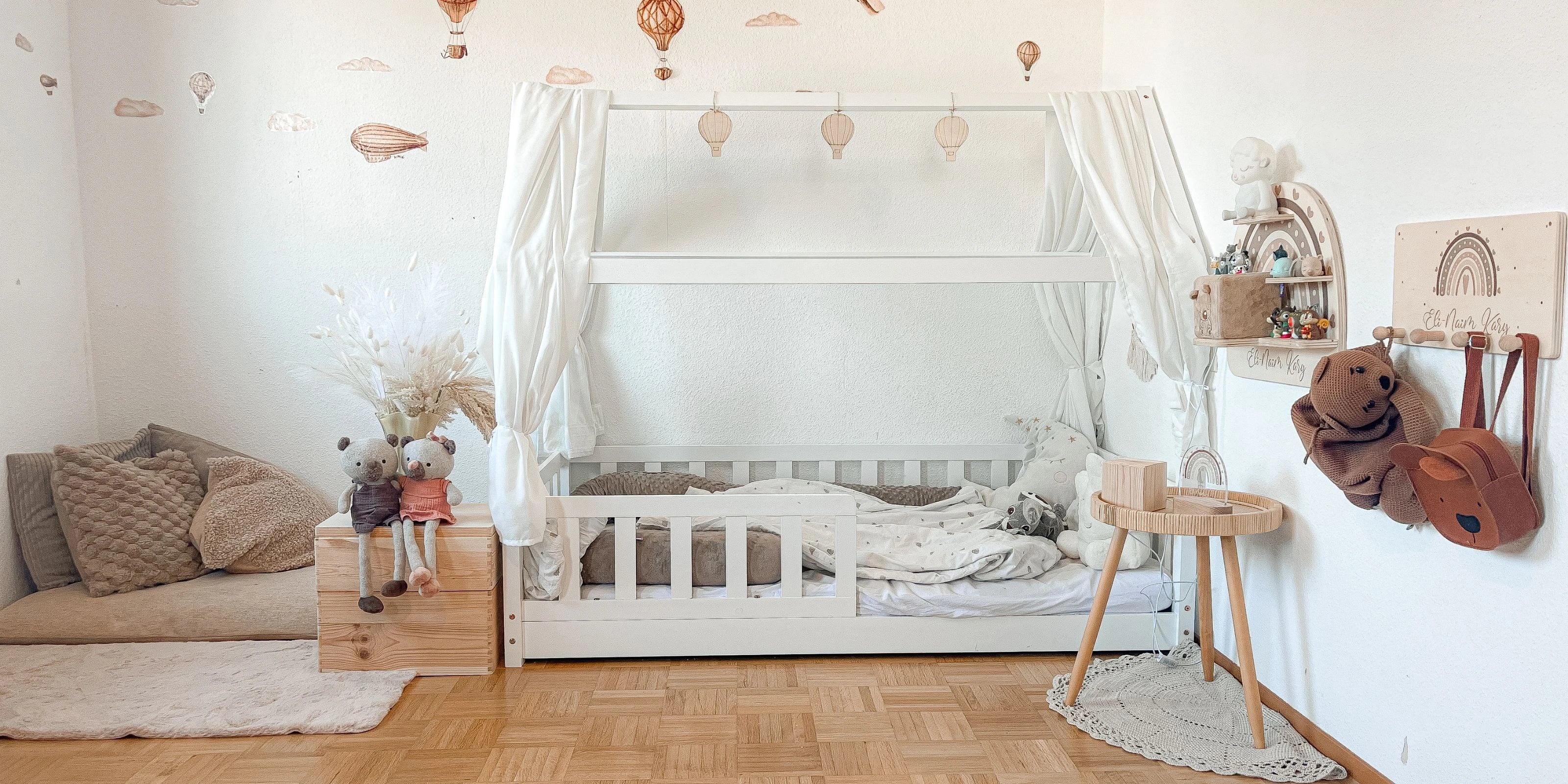 Gemütliches Kinderzimmer im Montessori-Stil mit weißem Hausbett, warmen Holzelementen und dekorativer Tapete mit Heißluftballons und Luftschiffen. Der helle Raum ist liebevoll mit Stofftieren, einem Beistelltisch und Wanddekoration eingerichtet - perfekt für erholsamen Schlaf und kreatives Spielen in kindgerechter Atmosphäre.