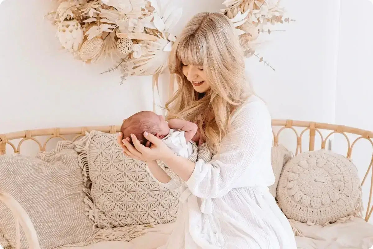Junge Mutter mit langen blonden Haaren in weißem Kleid hält liebevoll ihr neugeborenes Baby im Arm. Sie sitzt auf einer gemütlichen Rattanbank mit gestrickten Kissen in Naturtönen. Im Hintergrund hängt ein dezenter Trockenblumenkranz in neutralen Farben an der weißen Wand. Die Szene strahlt Wärme, Geborgenheit und den besonderen Moment der frühen Mutterschaft aus.